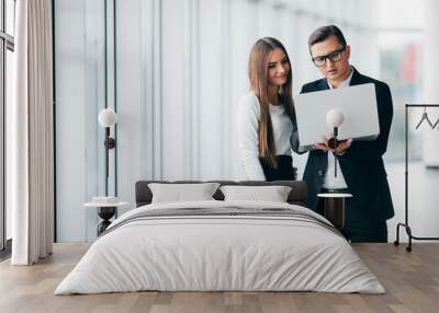 Businessman and businesswoman using a laptop together while standing in front of office building windows in office Wall mural